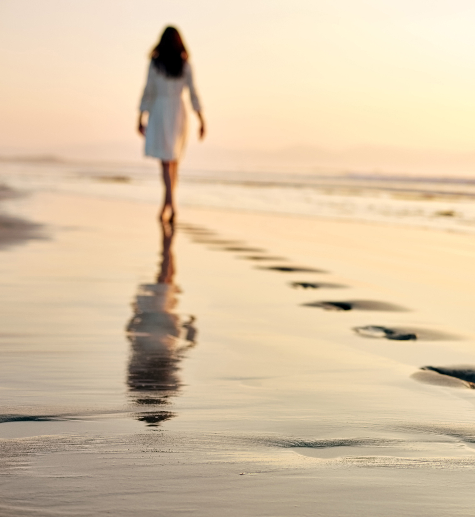 Woman walking along the beach shore leaving footprints in the sand at sunset